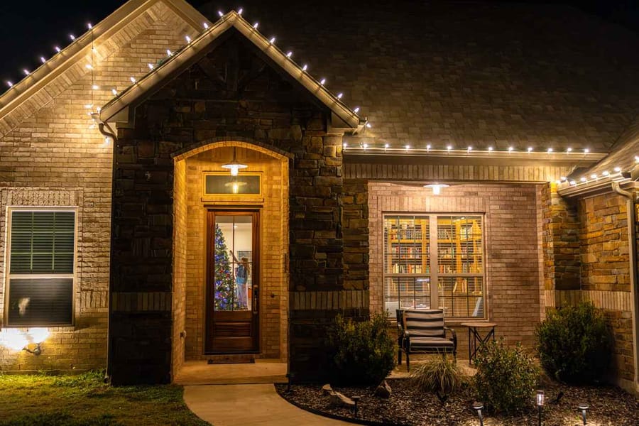 Home with outdoor holiday lighting and a young child decorating the Christmas tree through the window